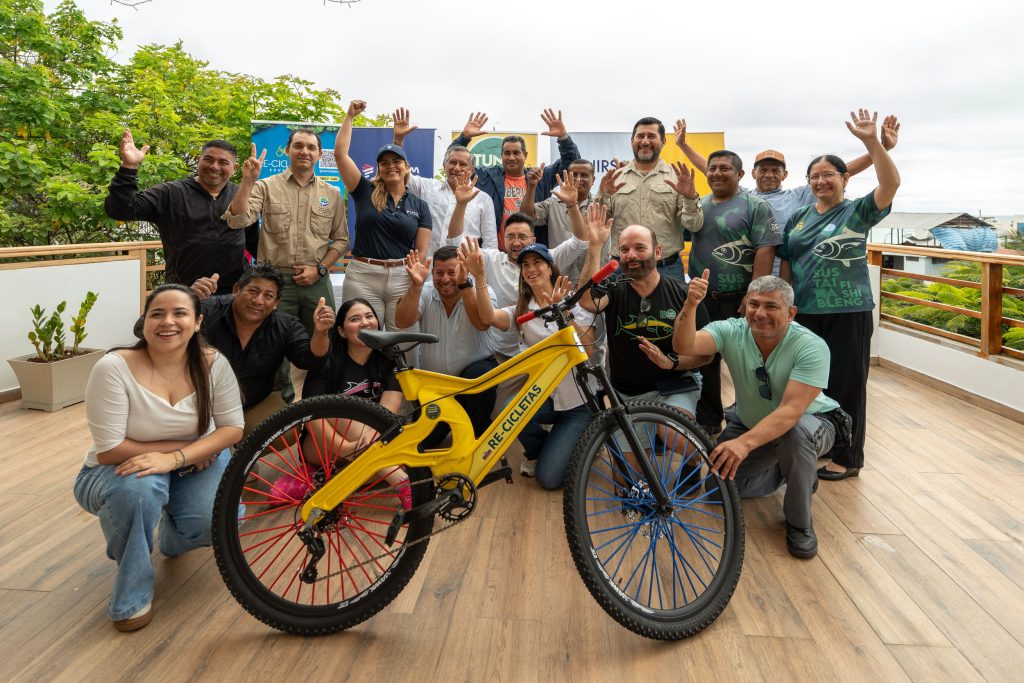 Iniciativa en Ecuador contra la contaminación marina donde colaboradores de NIRSA, Fundación Tunacons y pescadores artesanales posan con una bicicleta amarilla RE-CICLETAS hecha con plástico reciclado