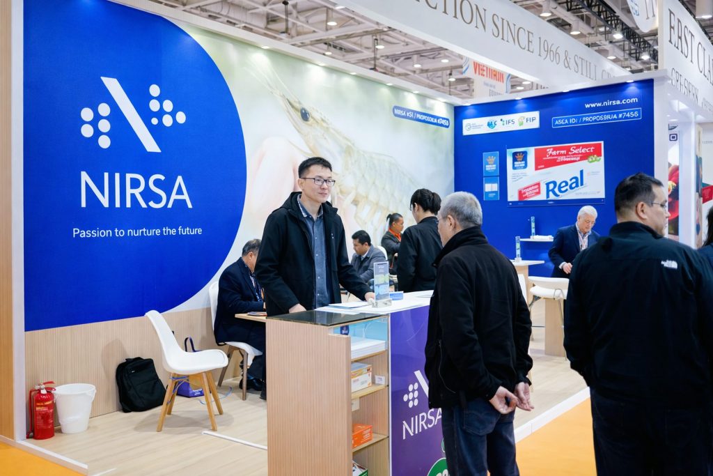 Stand de NIRSA en una feria: gran panel azul con el logo de la empresa, imagen de camarón y exhibición de productos Real, mientras visitantes conversan en el área de atención.